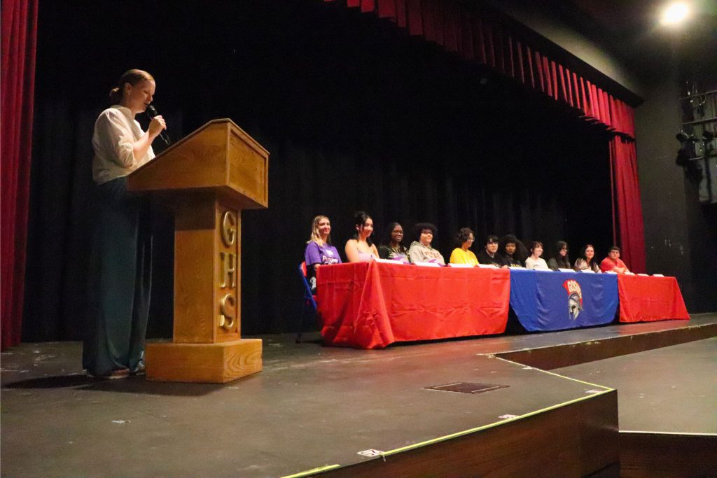 A side view of the stage, with a teacher talking at the podium and students sitting at the long red and blue table.