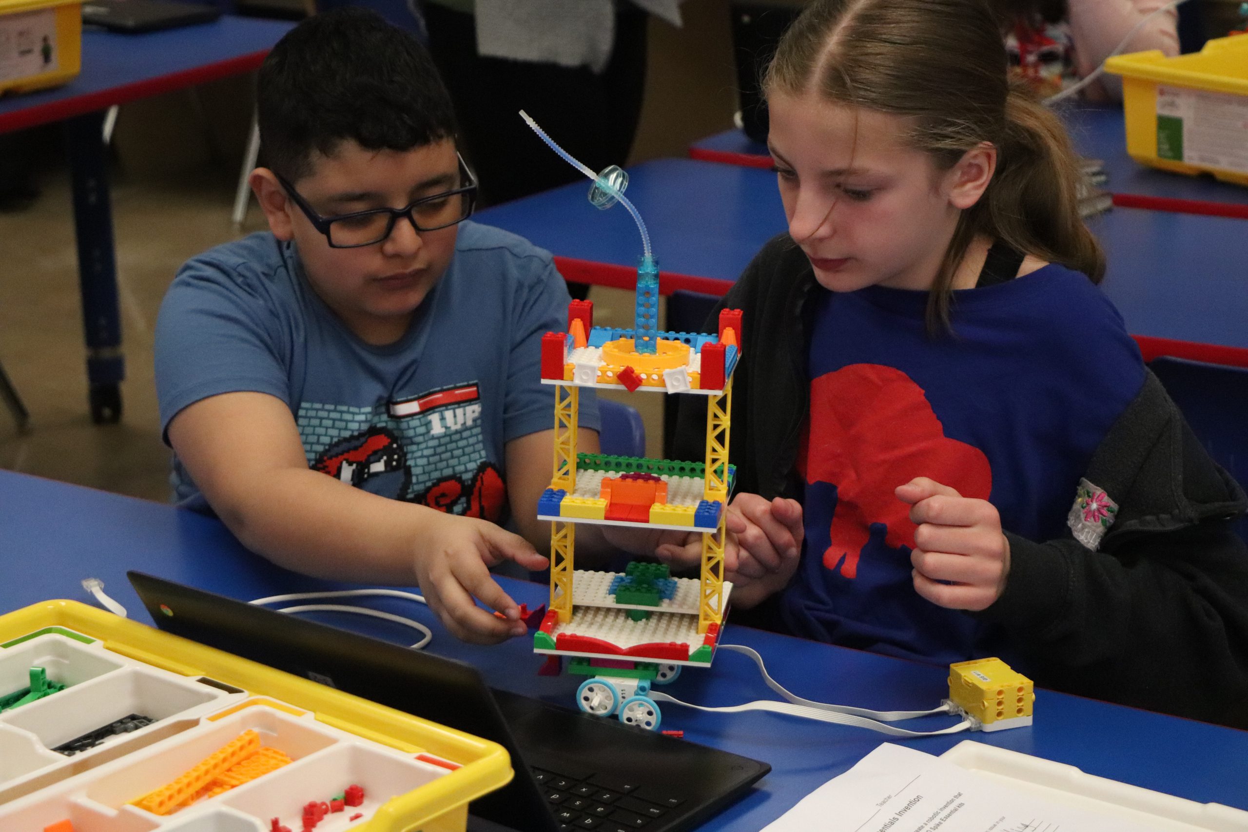 Two students are holding up a lego tower on wheels. 
