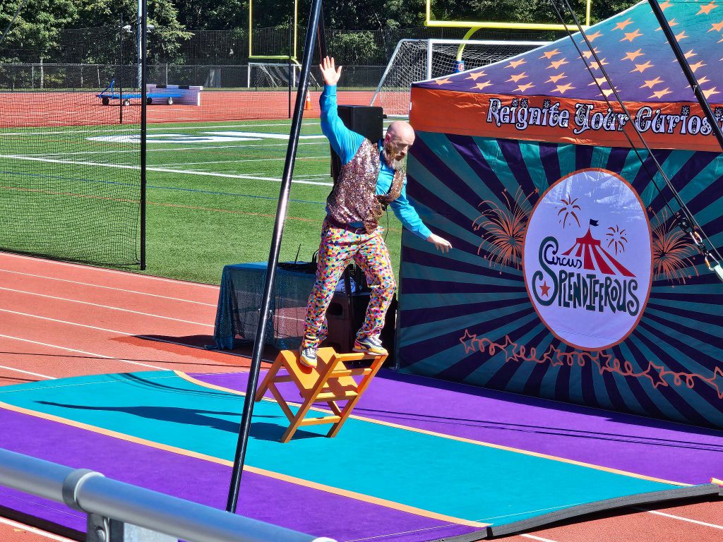 A performer leans a chair backward while standing on top of it. 