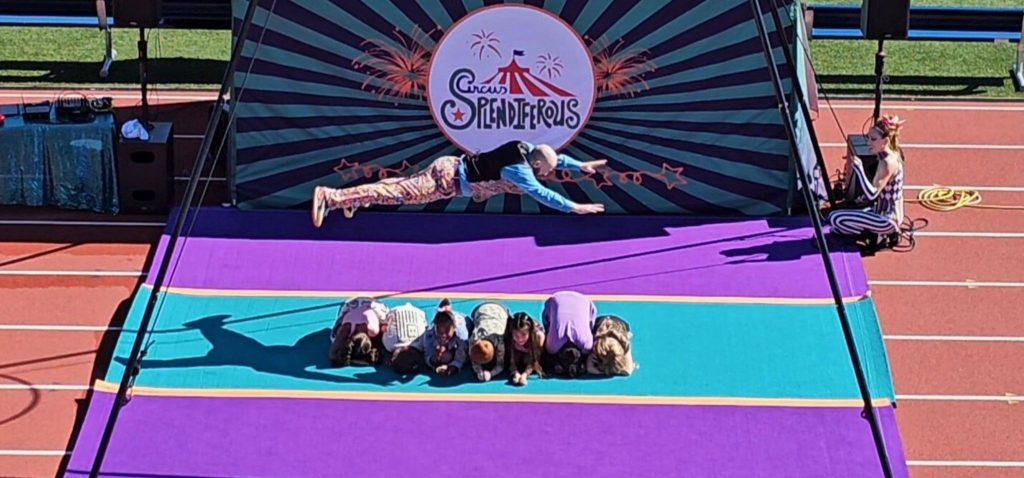 A performer jumps over students on the ground while fully extended in the air. The logo for Circus Splendiferous is behind them and a purple and blue matt is on the track. 