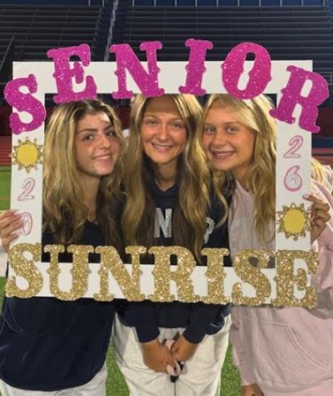 Three seniors at Goshen High School look through a senior sunrise 2026 cutout on the football field and smile for a photo.
