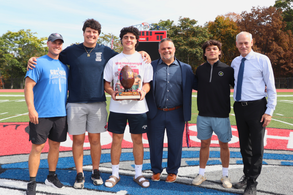 Goshen Football coach John Stephens, Senior Offensive Lineman Ryan, Senior Quarterback Jake Giordano, Goshen High School Principal Nick Pantaleone, Senior Running Back Liam O'Hara, Interim Athletic Director George St. Lawrence all stand for a photo and smile on the football field. Jake Giordano holds the Spirit Trophy. 