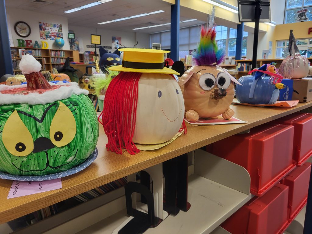 Pumpkins decorated as characters from books sit on the library shelves. Their faces are looking at the camera.