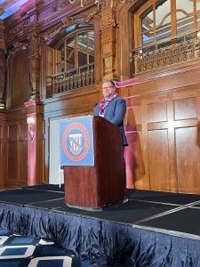 Adult stands on stage behind podium wearing a suit and tie.