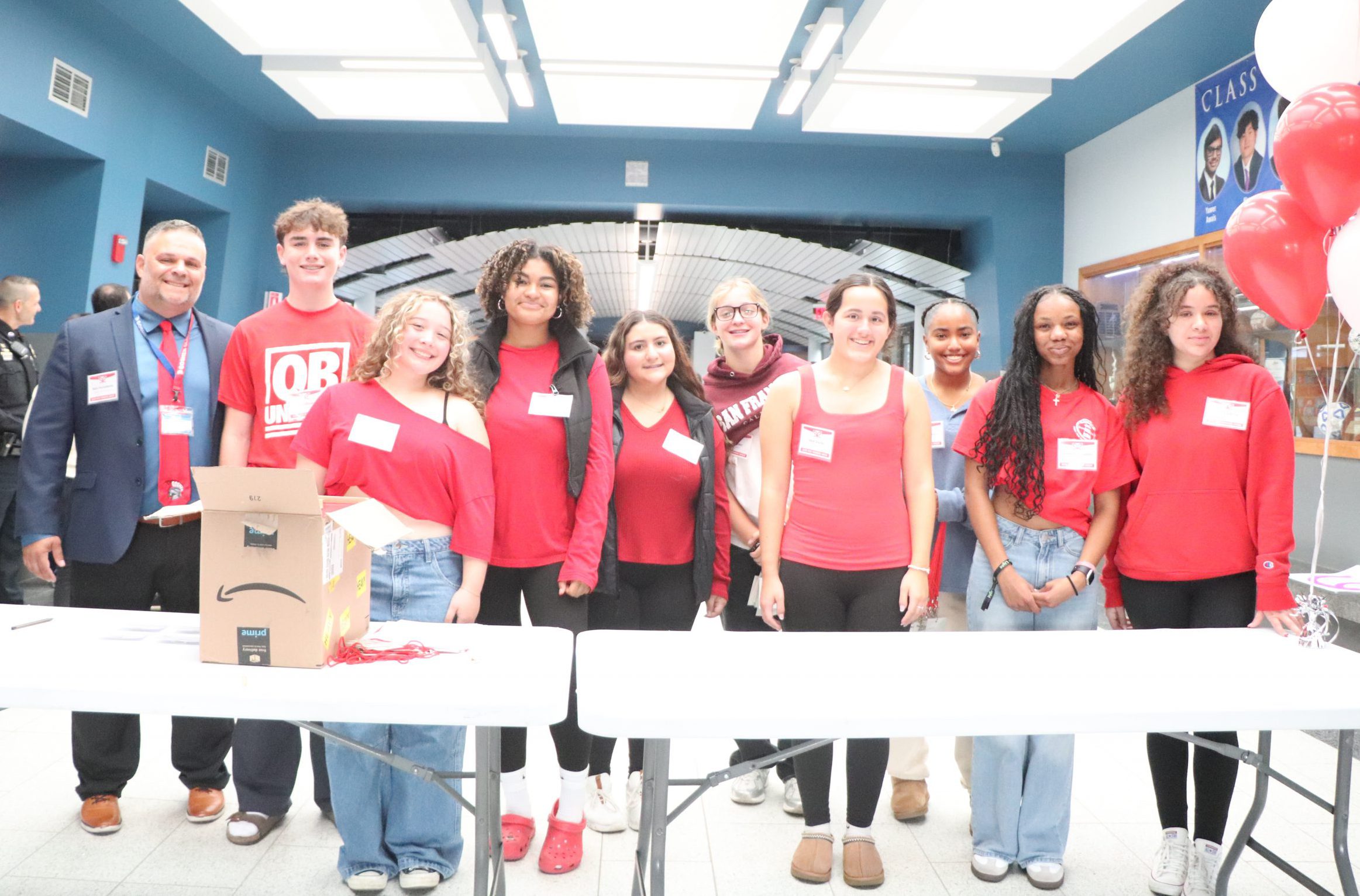 High school students wear red and stand behind a white table in the school lobby.