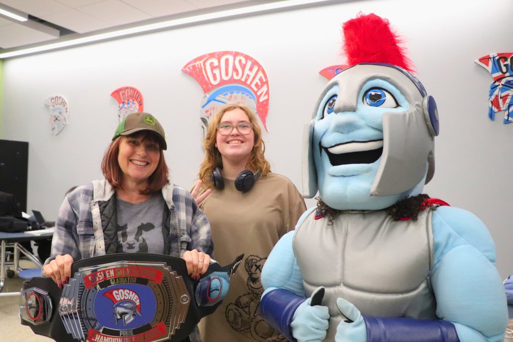 Mrs. Panzer stands on the left of the photo with the "Champion of Students" belt and smiles. A student is in the middle of the photo, and the Goshen Gladiator is on the right. The photo was taken in Goshen High School's cafeteria.