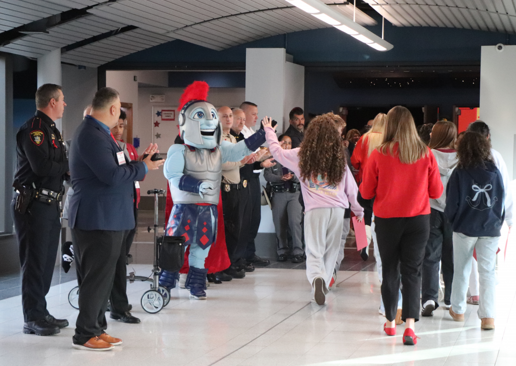 A mascot of a Gladiator with a grey and red helmet high fives students as they walk into a high school auditorium.