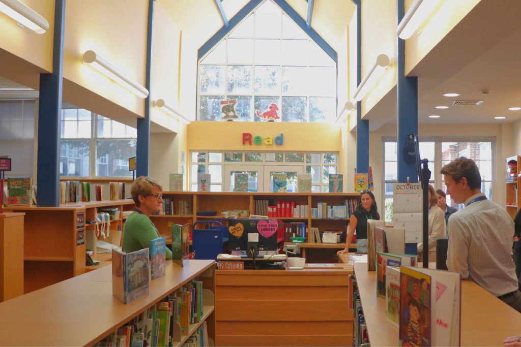 Amy Gargiulo speaks to a group of BOCES librarians in the Scotchtown Avenue Elementary School library. The sun is shining through the windows and there are books on the shelves.