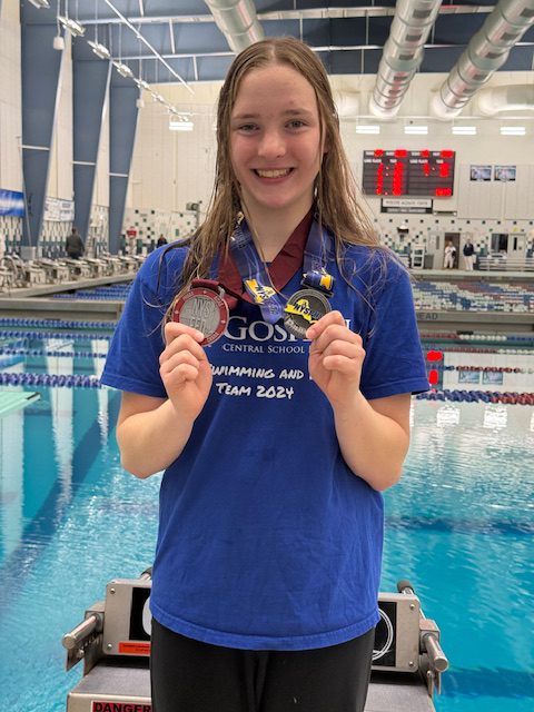 Teen with wet hair holds medals in front of a large swimming pool