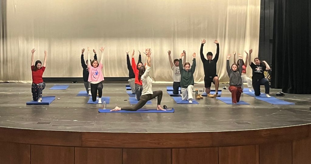 Teens on a high school auditorium stage performing yoga on blue yoga mats