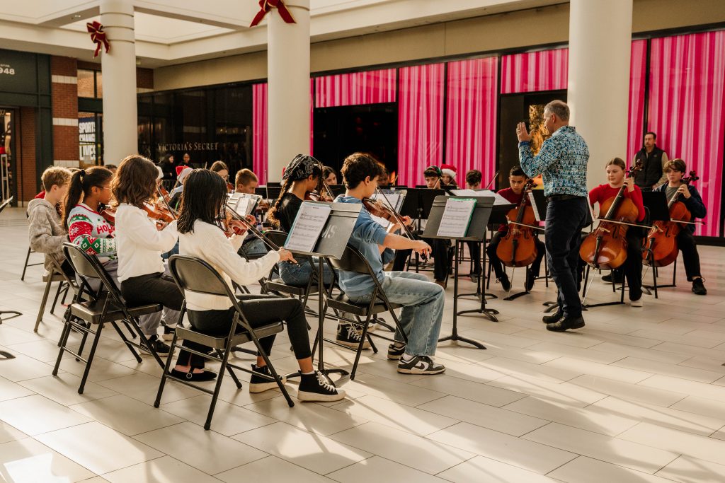 Orchestra students sit in folding chairs and perform in a mall