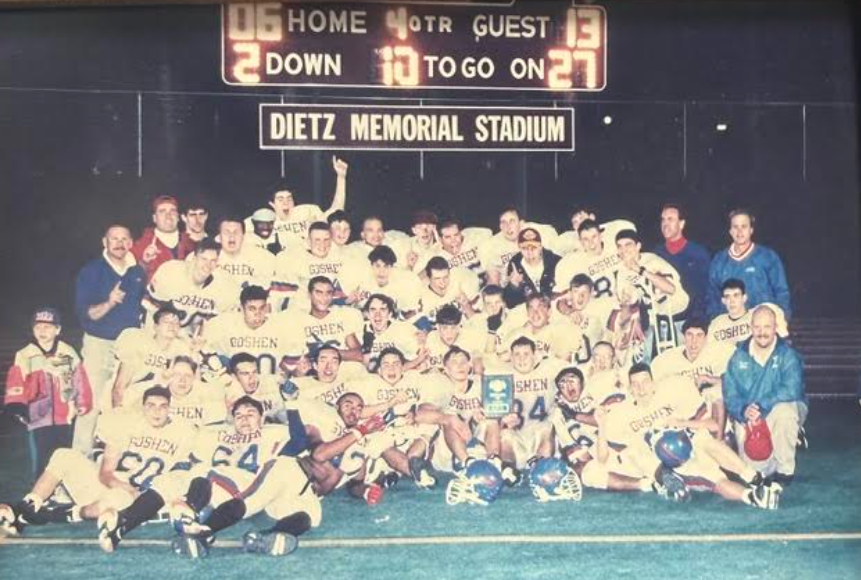 A faded photo of the 1994 football team wearing white jerseys with blue letters and their coaches; all are cheering.
